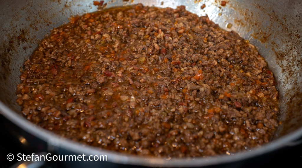 A pan filled with a rich and savory meat ragù, featuring finely chopped meat and vegetables in a dark sauce, simmering on a stovetop.