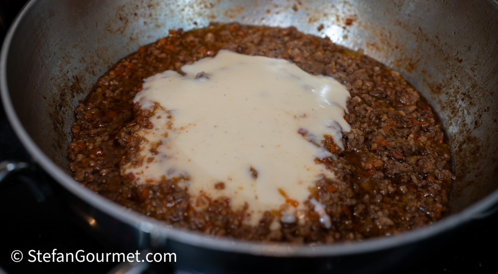 A close-up view of a pan containing a meat ragù mixture with a layer of creamy besciamella sauce on top, ready to be stirred together.