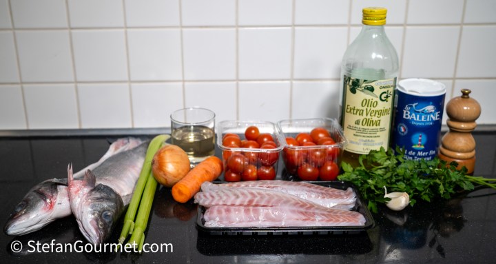 Ingredients for Branzino all'Acqua Pazza including whole branzino, branzino fillets, cherry tomatoes, onion, carrot, celery, garlic, olive oil, white wine, and seasonings on a kitchen countertop.