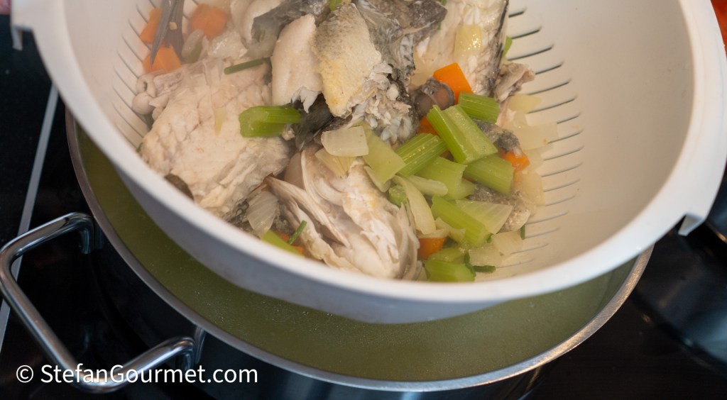 Strainer filled with fish heads and assorted vegetables, resembling ingredients for making fish stock.