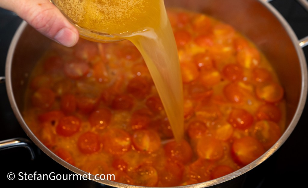 A hand pouring fish stock into a pot of halved cherry tomatoes, simmering on the stove.