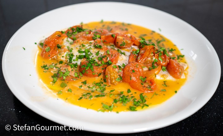A plated dish of poached branzino topped with cherry tomatoes and fresh parsley in a light broth.