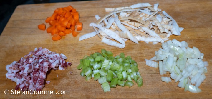 Chopped ingredients for cooking, including diced carrots, parsley root, celery, onion, and pancetta arranged on a wooden cutting board.