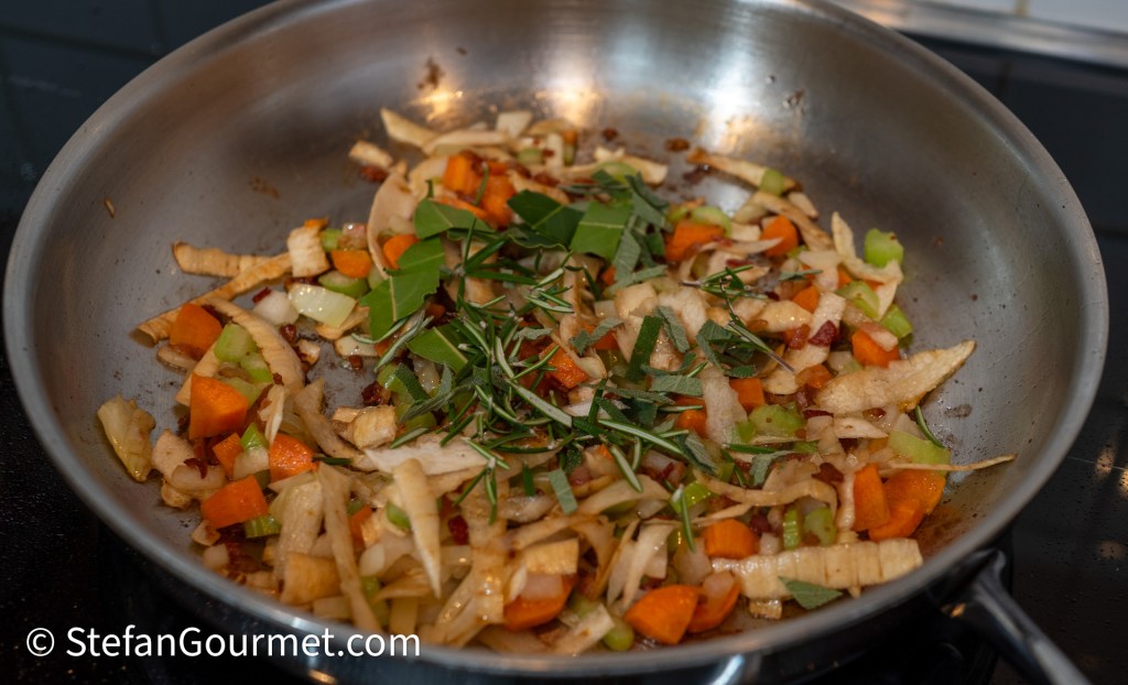 A pan filled with sautéed vegetables including carrots, celery, and onion, along with herbs like rosemary and bay leaves, all slightly caramelizing on a stovetop.