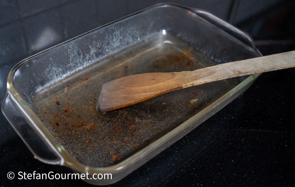 An empty glass dish with remnants of a roasted meat preparation and a wooden spatula resting inside.