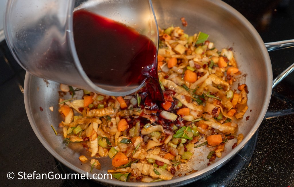 A stream of red wine being poured into a stainless steel skillet containing sautéed vegetables, including diced carrots, celery, and parsley root.