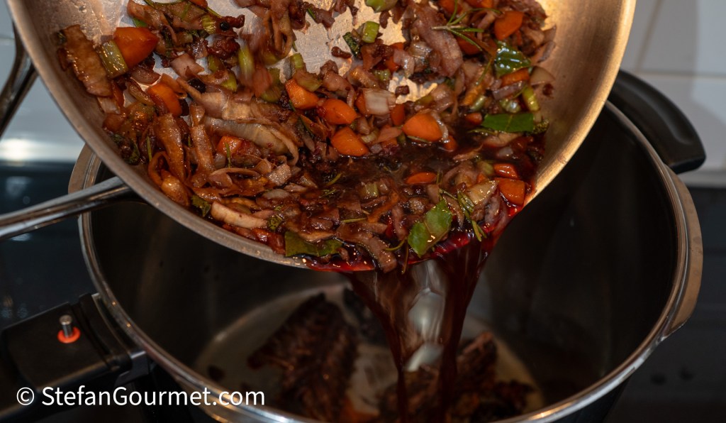 A saucepan pouring sautéed vegetables and red wine into a pressure cooker, showcasing the cooking process for a rich sauce.