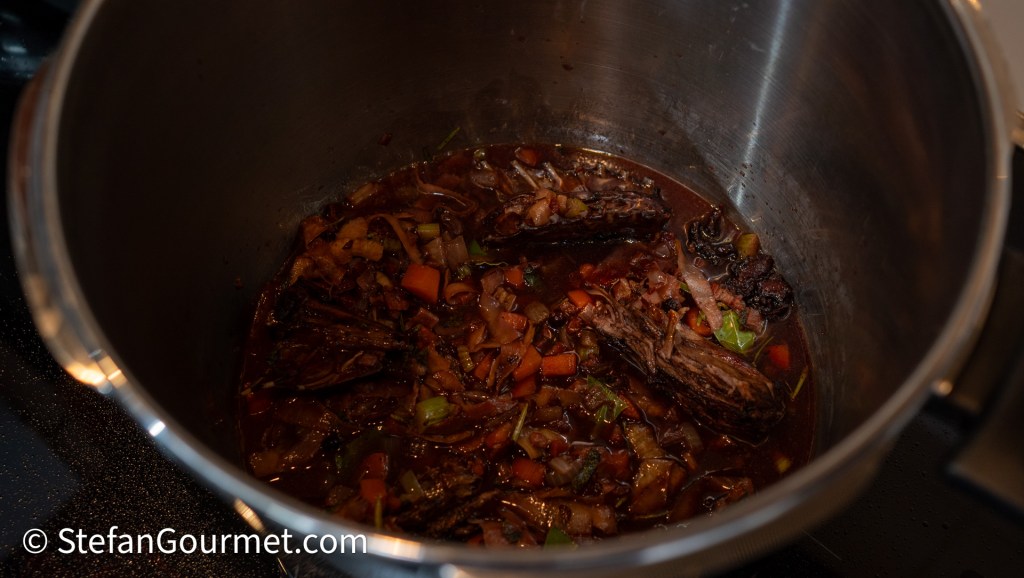 A stainless steel pot filled with simmering hare stock, chunks of vegetables, and bones visible in the rich, dark liquid, reflecting the process of making a complex sauce.