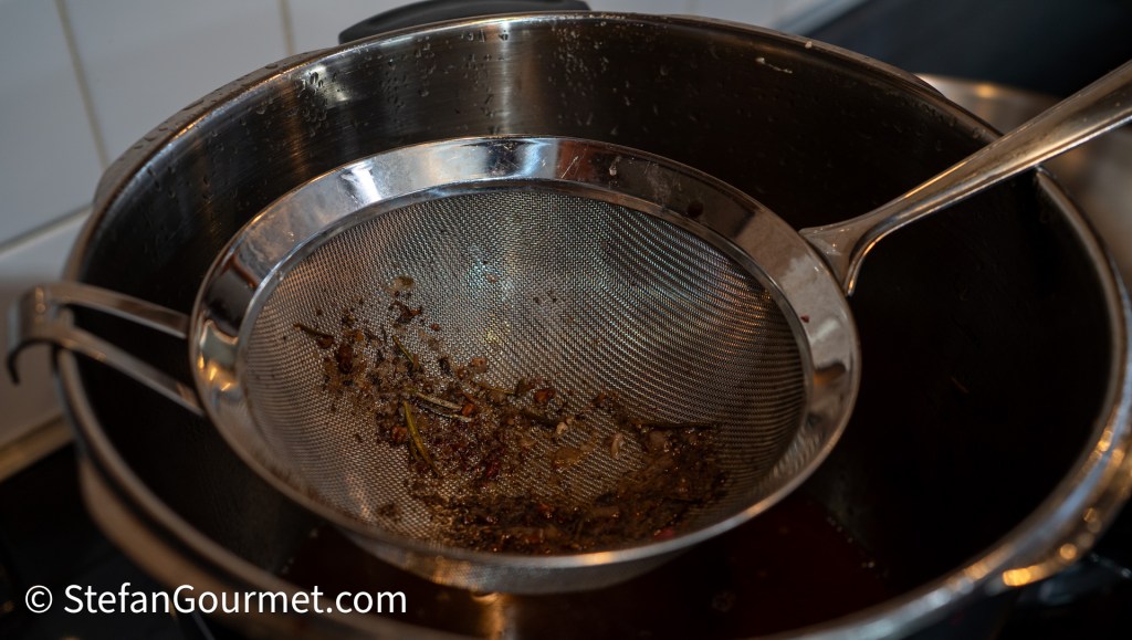A fine mesh strainer over a pot, containing strained remnants of herbs and spices used for a sauce, with steam rising.