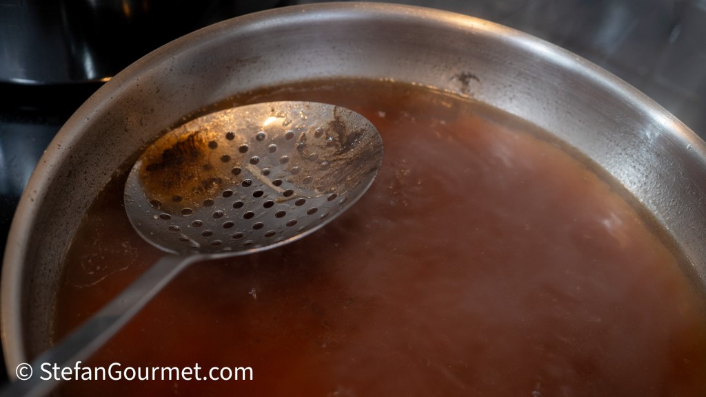 A close-up of a stainless steel pot simmering with a rich brown sauce, featuring a slotted spoon resting on the edge.