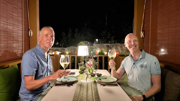 Two men seated at a elegantly set dinner table in a romantic outdoor dining setting at night, raising glasses of wine in a toast.