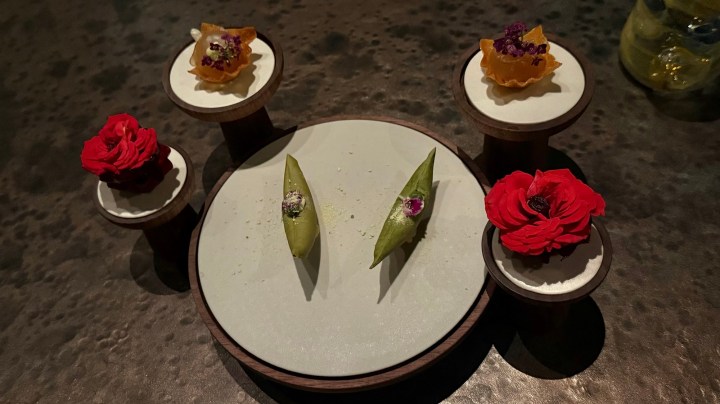 A beautifully presented dish of amuse-bouches featuring edible flowers, garnishes, and green pea pods on a round white plate, set on wooden stands.