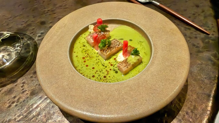 A dish featuring pieces of kingfish resting in a vibrant green sauce, garnished with red elements and fresh herbs, served in a round stone plate.