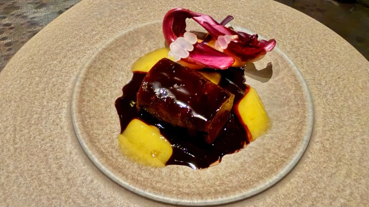 A beautifully presented dish featuring a rectangular piece of meat in a rich dark sauce, accompanied by slices of yellow fruit and garnished with vibrant pink edible flowers on a round beige plate.