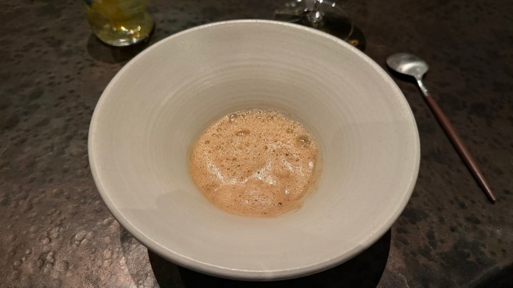 A shallow white bowl containing a frothy, bubbling liquid, placed on a textured dark surface.
