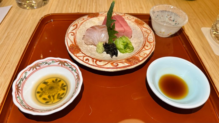 A plate of sashimi featuring sea bream and tuna, garnished with wasabi and seaweed, served with dipping sauces in small bowls on a wooden tray.