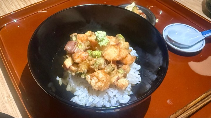 A bowl of rice topped with mixed tempura, featuring various vegetables and seafood, served on a wooden tray.