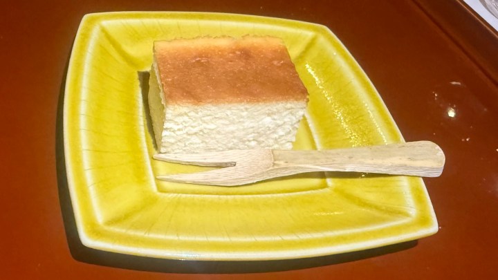 A slice of baked cheesecake served on a yellow ceramic plate, accompanied by a wooden fork.