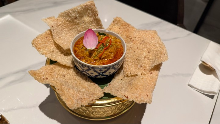 A bowl of crab curry dip served with crispy rice crackers, garnished with a pink lotus petal and thin slices of chili.