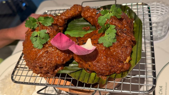 Grilled chicken legs marinated in a thick curry sauce, garnished with cilantro and a pink lotus flower petal, served on a wire rack with banana leaves.