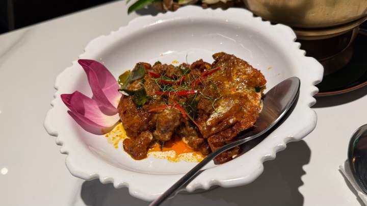 A serving of a traditional Thai dish in a white bowl, featuring a rich curry and garnished with herbs, red chilies, and decorative lotus petals.