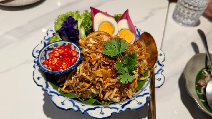 A vibrant plate featuring Thai-style Lotus Flower Salad, garnished with crispy fried shallots, fresh herbs, cherry tomatoes, boiled egg slices, and a side of red chilies.