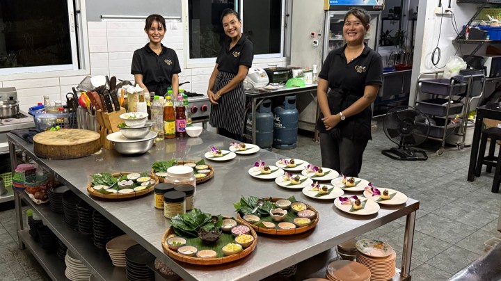 Three women in black uniforms standing in a restaurant kitchen, smiling and posing for the camera. The kitchen counter is adorned with various prepared dishes and ingredients.
