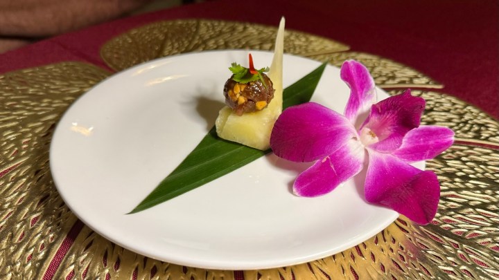 A beautifully presented amuse-bouche featuring a slice of fresh, sweet pineapple topped with a rich pork and peanut meatball, garnished with herbs and a vibrant pink orchid flower.