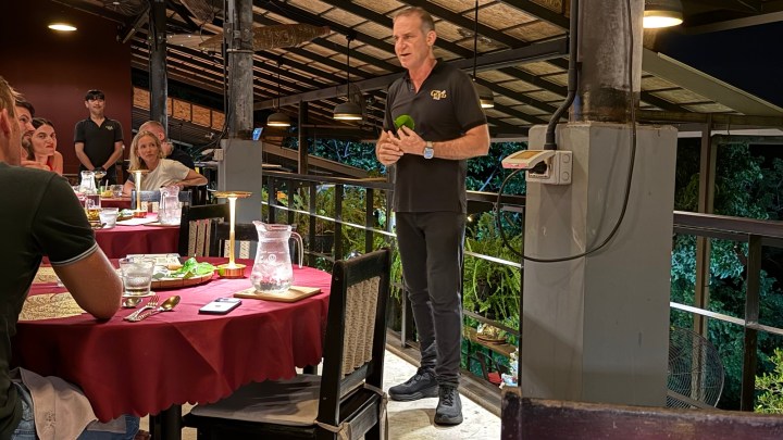 A chef explaining a dish to guests at a restaurant with a view, surrounded by tables set for dining.