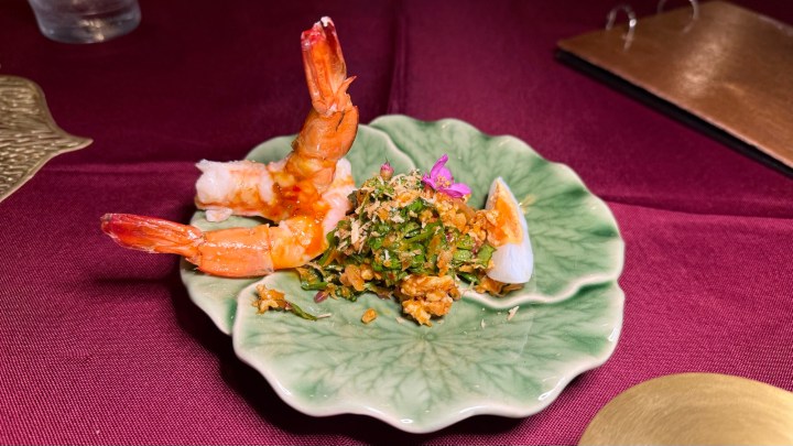 A green plate with two large prawns and a pile of mixed herbs and crunchy toppings, garnished with pink and purple flowers.