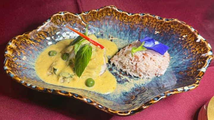 A beautifully plated Thai dish featuring a piece of fish in a creamy green curry sauce, garnished with a red chili and fresh basil leaves, served alongside a portion of multi-colored rice with a purple flower garnish.