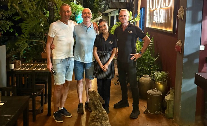 A group photo of four people, including restaurant staff, taken in front of the Gold Bar sign, with a leafy background.
