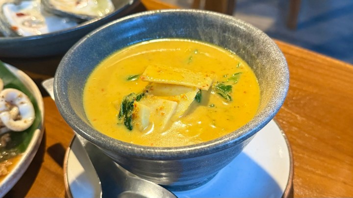 A bowl of Thai coconut curry with tofu and greens, served with a spoon on a wooden table.