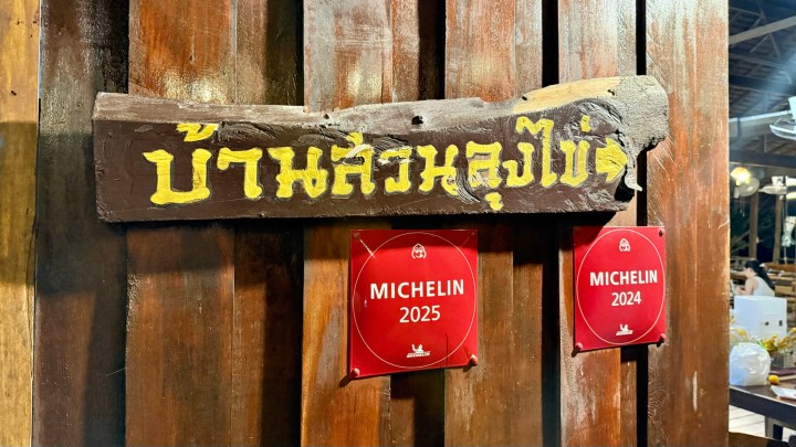 A wooden sign with Thai writing indicating 'Ban Suan Lung Khai,' alongside two red Michelin award plaques for 2024 and 2025, mounted on a wooden wall.