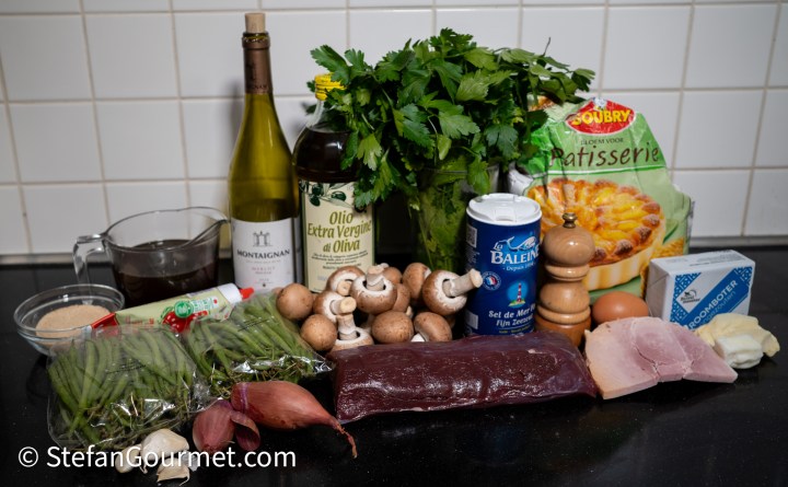 A collection of cooking ingredients including wine, olive oil, fresh parsley, mushrooms, green beans, shallots, a piece of meat, eggs, butter, and a pastry mix on a kitchen countertop.