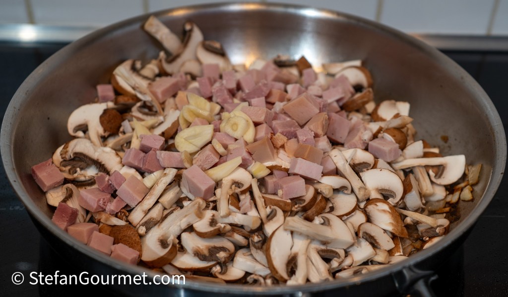 A frying pan filled with sliced mushrooms and diced pieces of ham, along with chopped garlic.