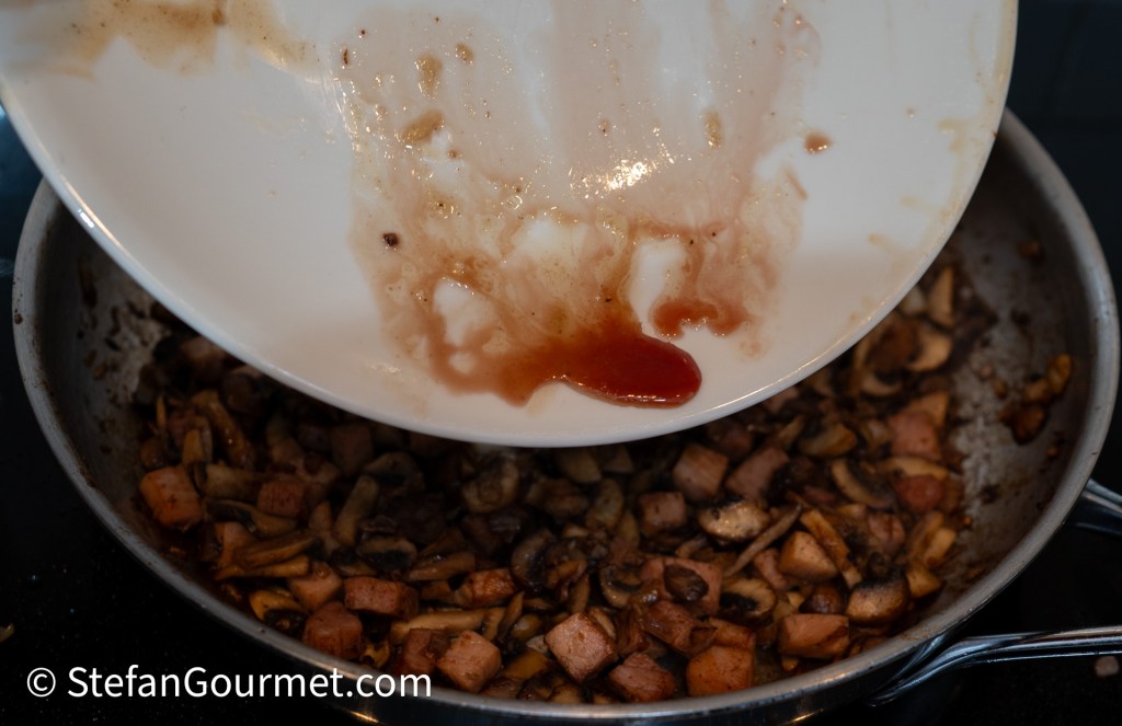 A bowl being tilted over a skillet with sautéed mushrooms and diced ingredients, with residue sauce dripping from the bowl.