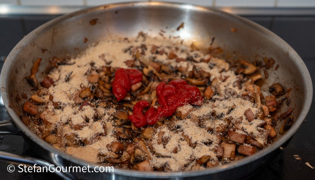 A stainless steel skillet containing sautéed mushrooms, diced bacon, and a layer of breadcrumbs, with blobs of tomato paste in the center.