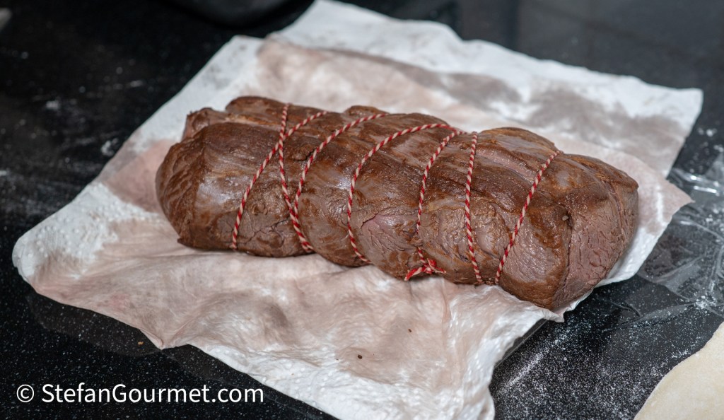 A tied piece of meat resting on parchment paper on a black countertop.