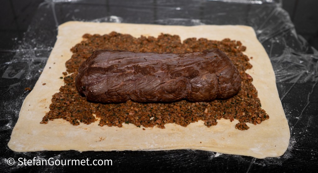 A raw piece of beef placed on a layer of finely chopped herbs and spices, surrounded by a sheet of pastry on a countertop.