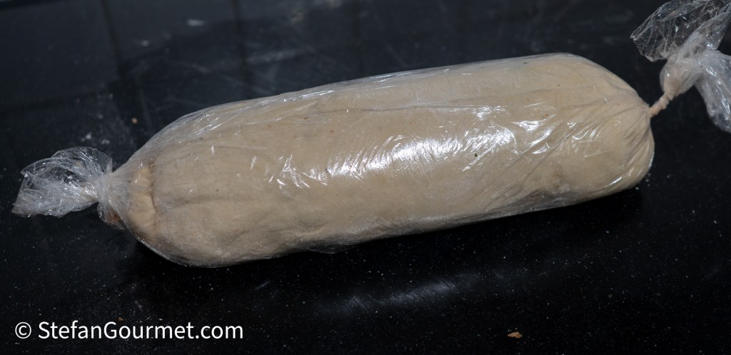 A wrapped cylinder of dough or paste stored in plastic film, resting on a dark countertop.