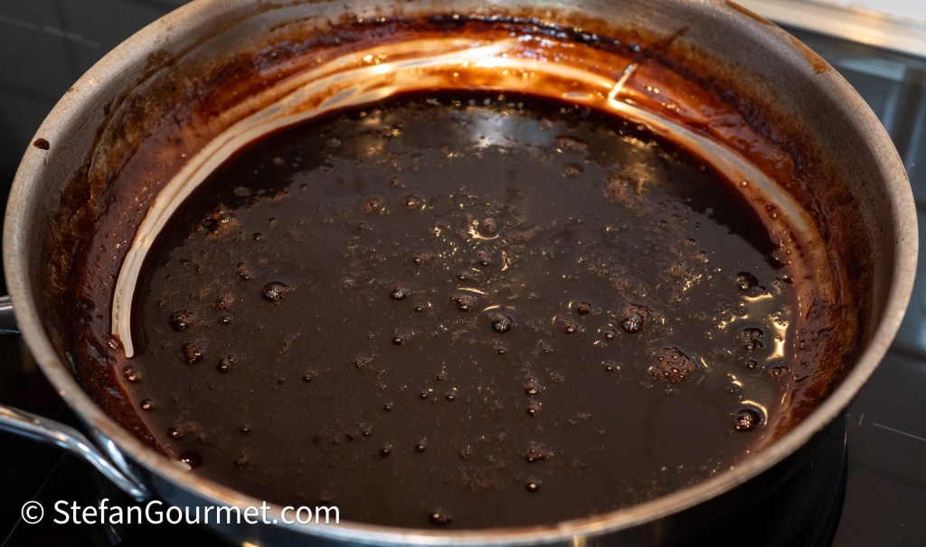 A stainless steel pan containing a dark, syrupy sauce with bubbles, indicating it is being cooked or reduced.