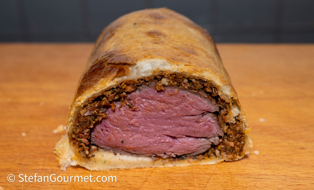 Cross-section of a well-cooked beef Wellington, showcasing the tender pink meat surrounded by a layer of ground mushrooms and herbs, all wrapped in a golden pastry crust.