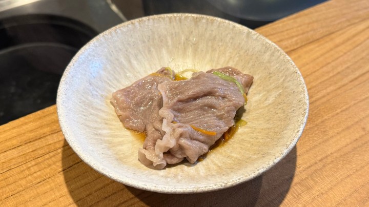 A small bowl containing sliced raw meat with a garnish of vegetables, resting on a wooden surface.