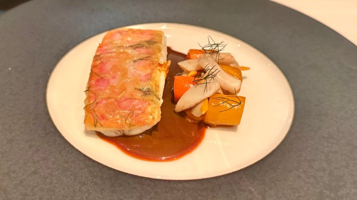 A plated dish featuring a portion of fish topped with a colorful layer, accompanied by diced squash and sliced mushrooms in a savory sauce.