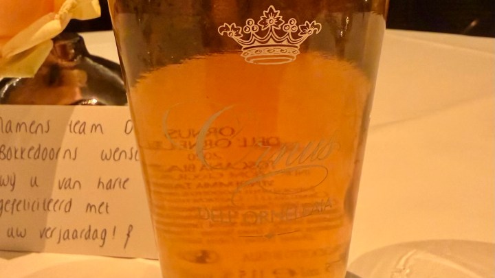 A glass containing a golden beverage with a decorative crown logo, sitting on a table alongside a note.