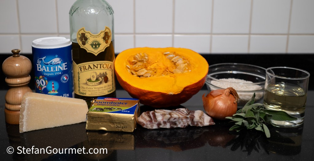 An assortment of cooking ingredients displayed on a countertop, including a half pumpkin, olive oil, rice, a shallot, fresh sage, bacon, grated cheese, salt, and a pepper mill.