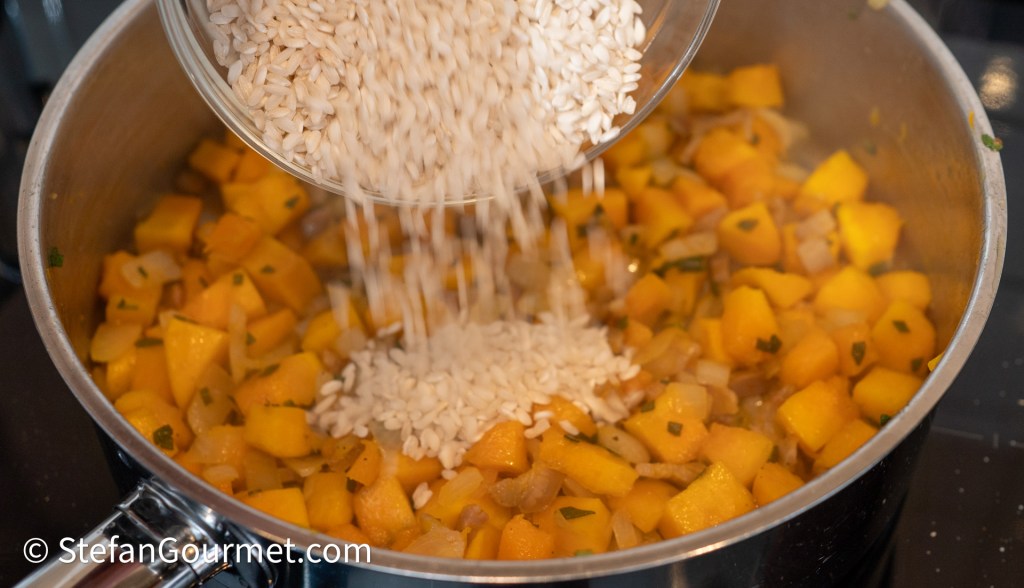 A pot on a stove with diced butternut squash and onions, as rice is being poured into it.