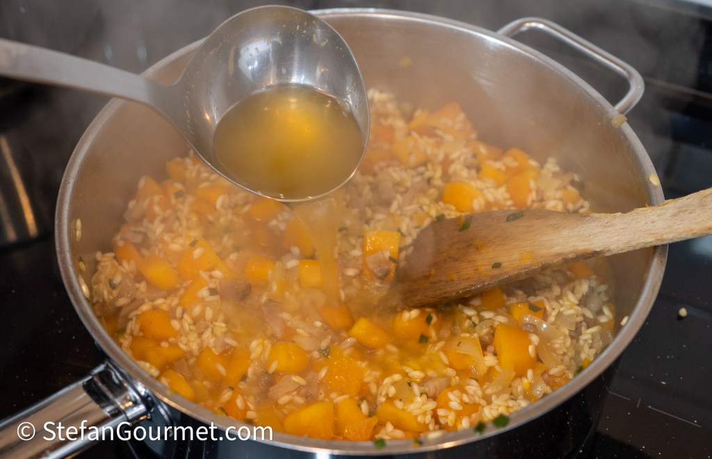 A saucepan containing risotto with diced butternut squash is being stirred. A ladle is pouring broth into the mix, creating steam.