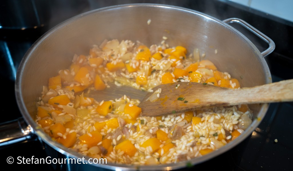 A pot containing simmering risotto with diced pumpkin and onions, being stirred with a wooden spoon.